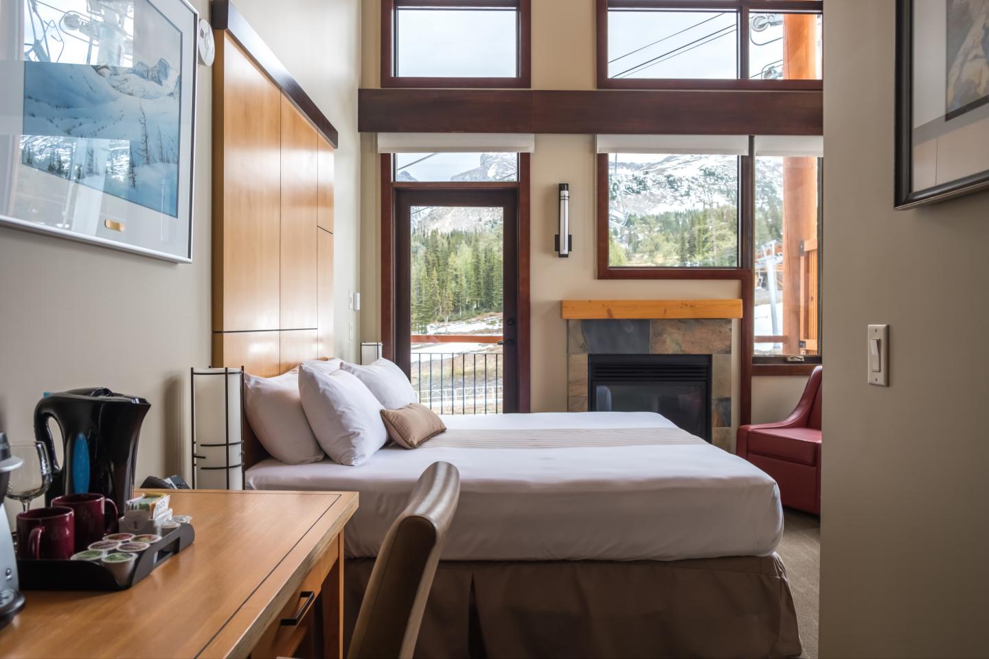 Modern hotel room with large bed, fireplace, and desk. Bright windows overlook greenery.