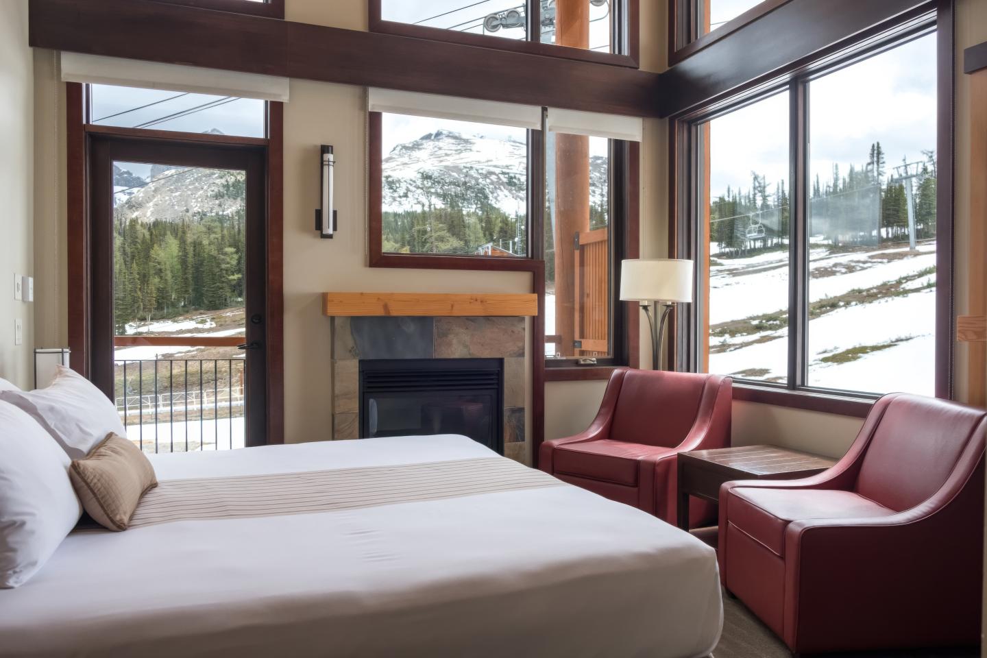 Cozy bedroom with mountain view through large windows, red chairs, and a fireplace.