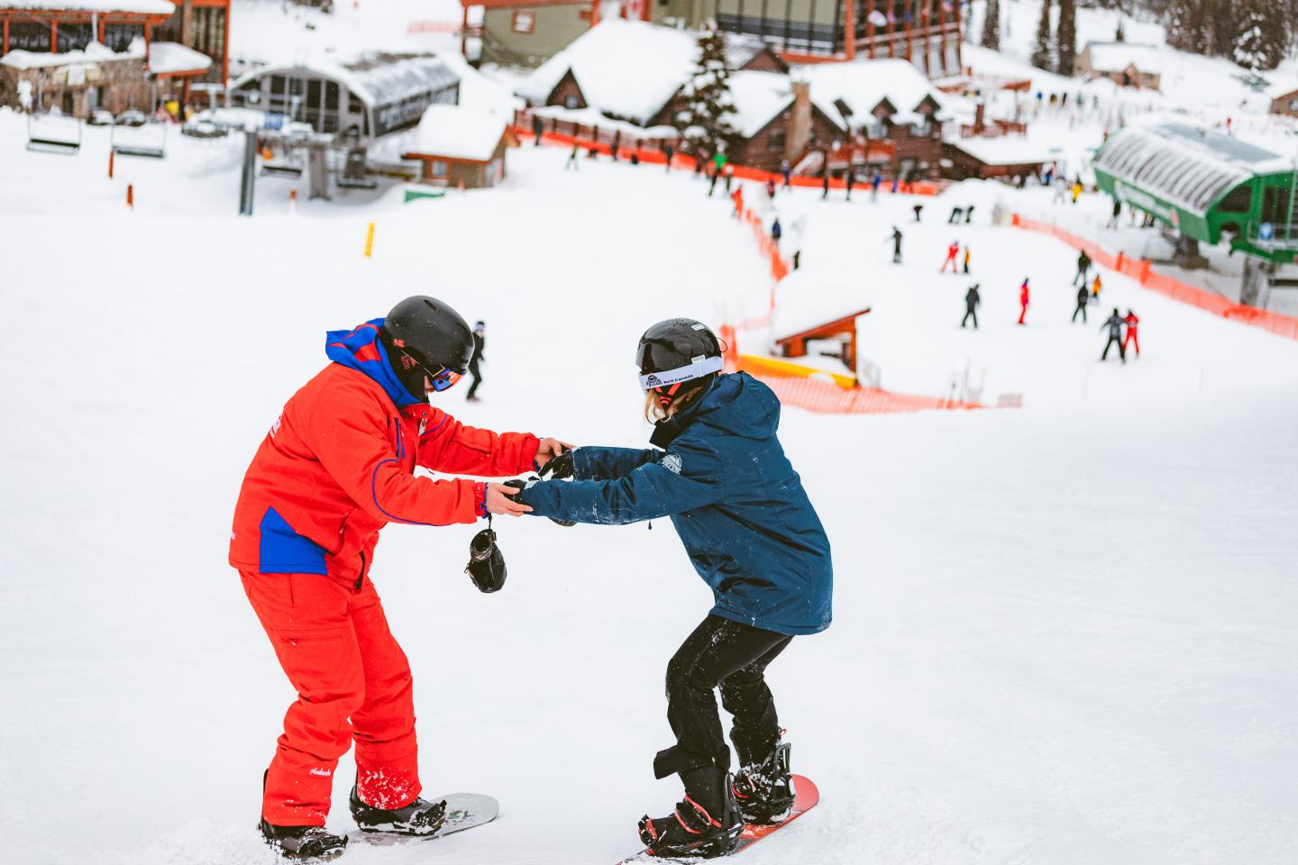 Two snowboarders in red and blue jackets holding hands on a snowy slope.