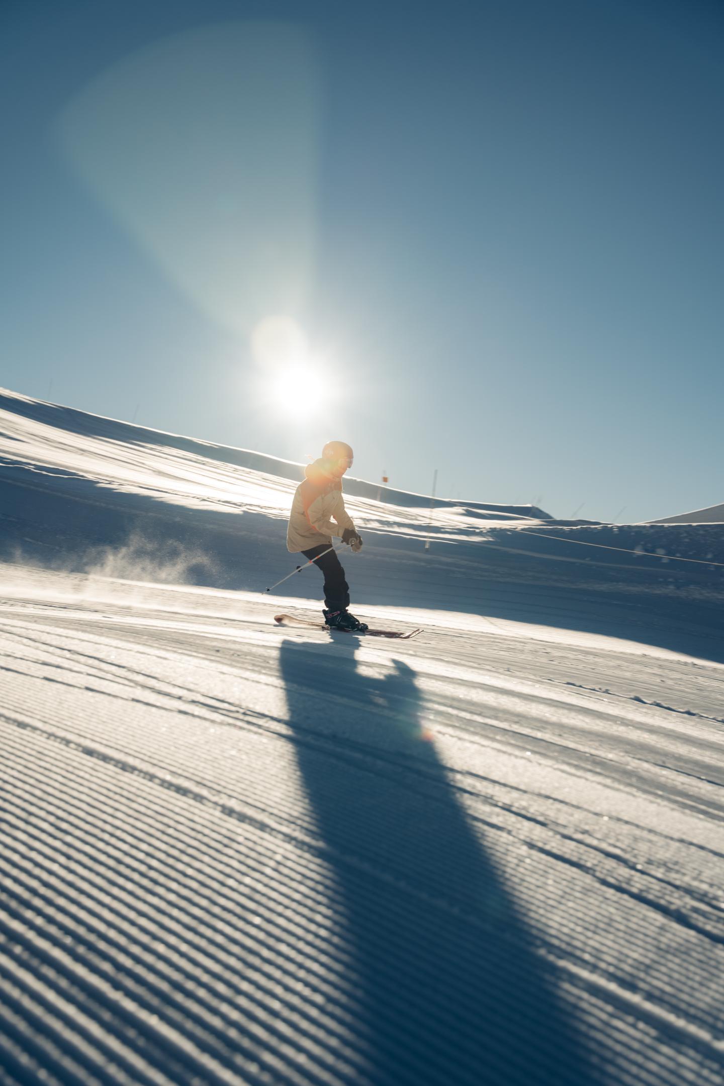 Snowboarder standing on a sunlit, groomed slope, casting a long shadow.