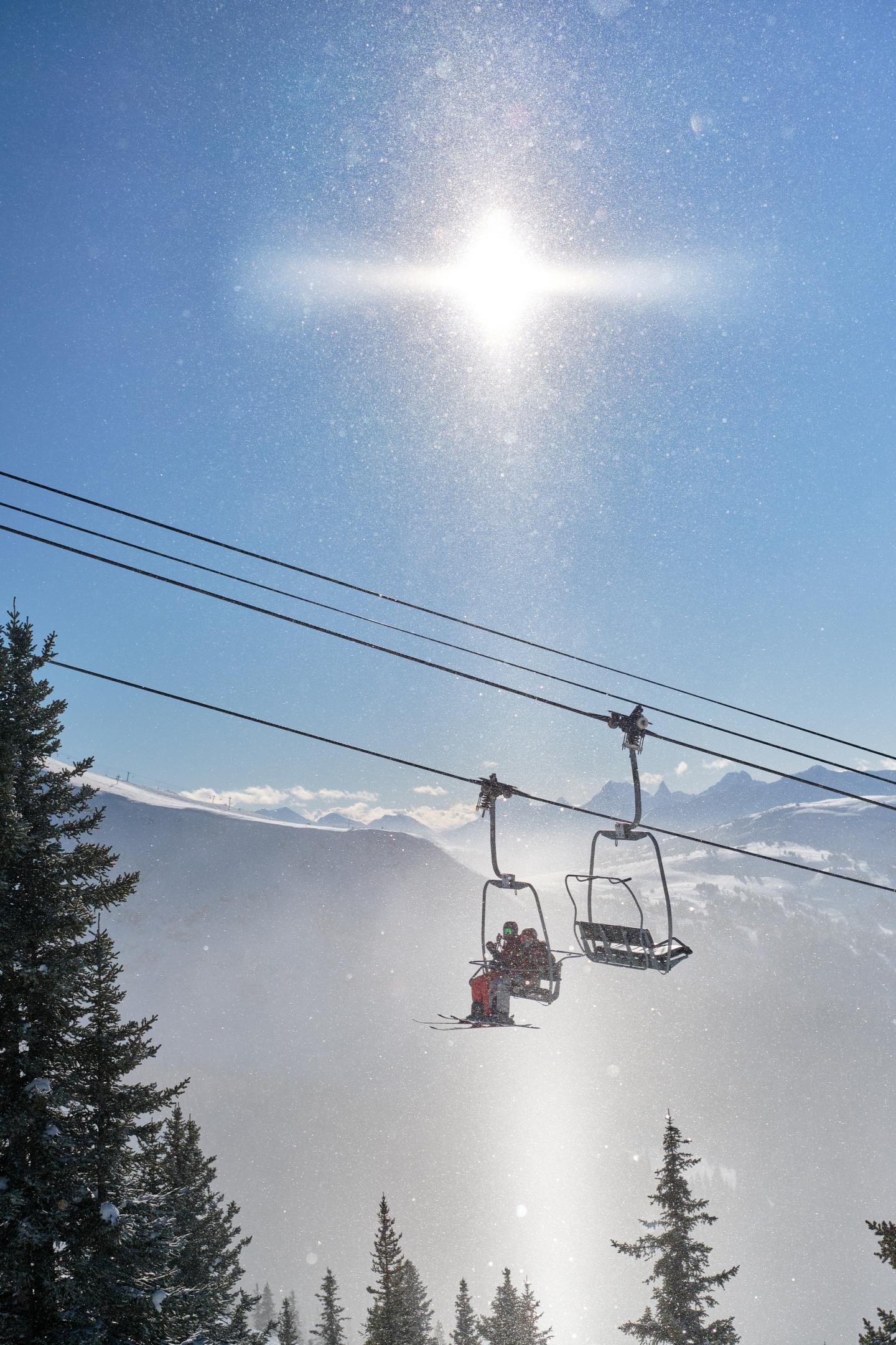 Sun shining over a snowy mountain ski lift and pine trees.