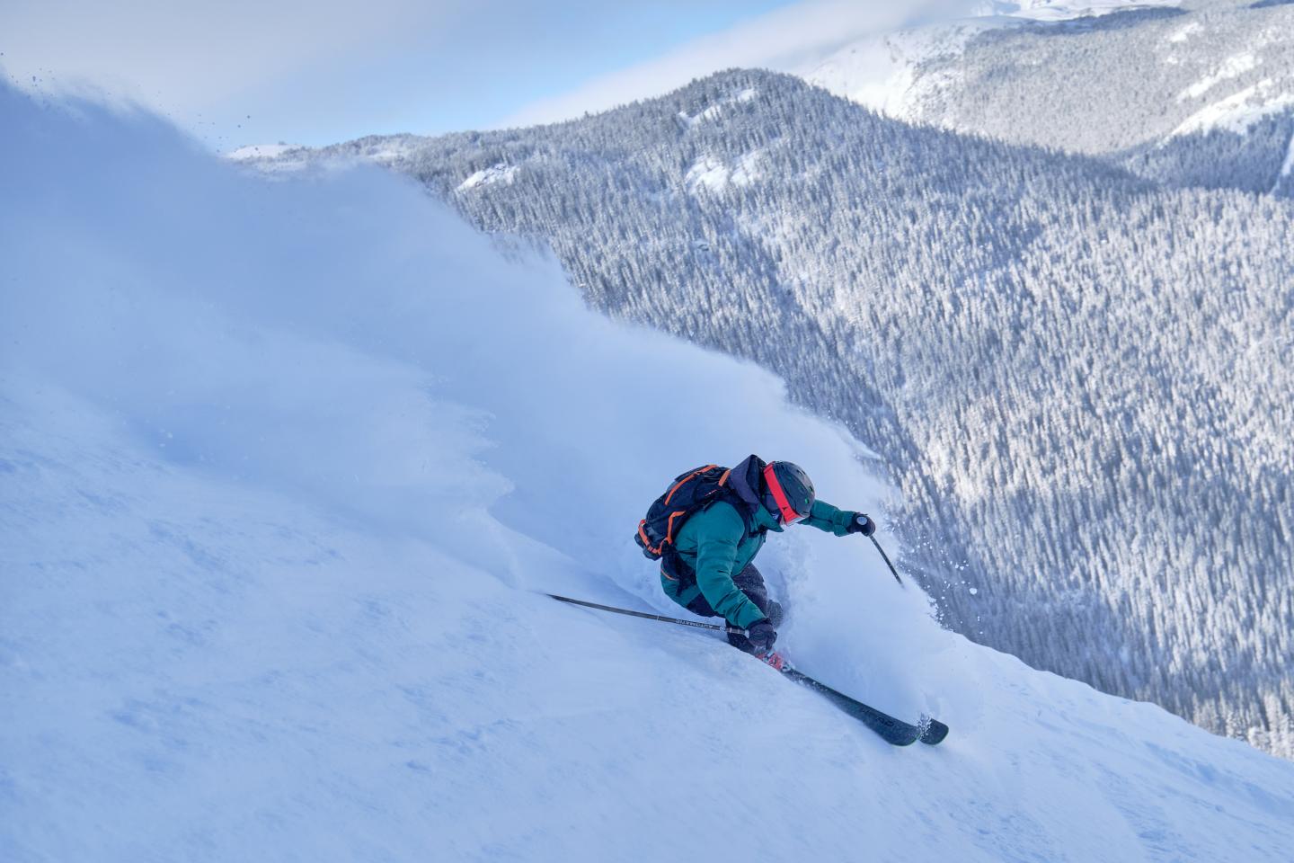 Skier carving down a steep, snowy mountain slope beneath forested peaks.