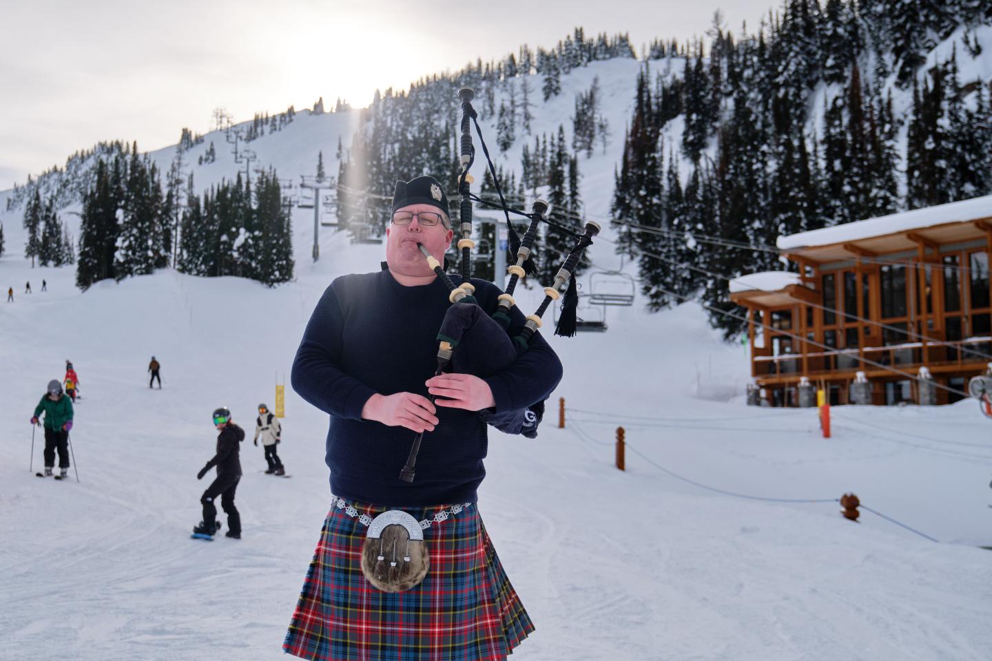 Man in tartan kilt plays bagpipes on a snowy ski slope.