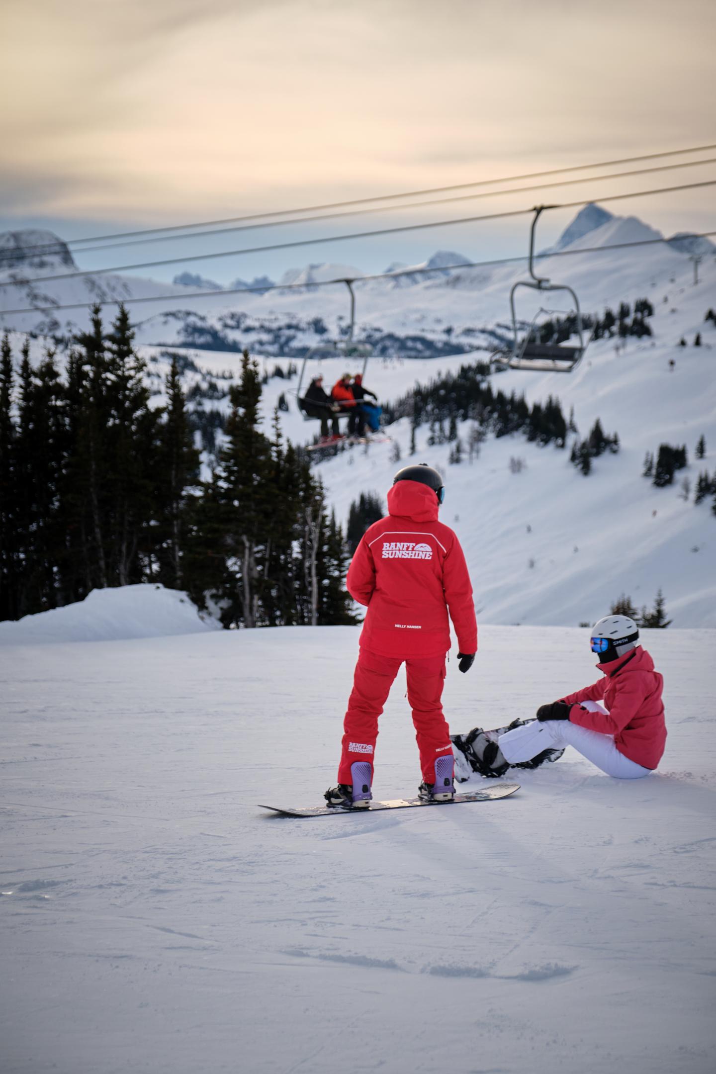 Two skiers in red outfits on a snowy slope overlooking a mountain valley.