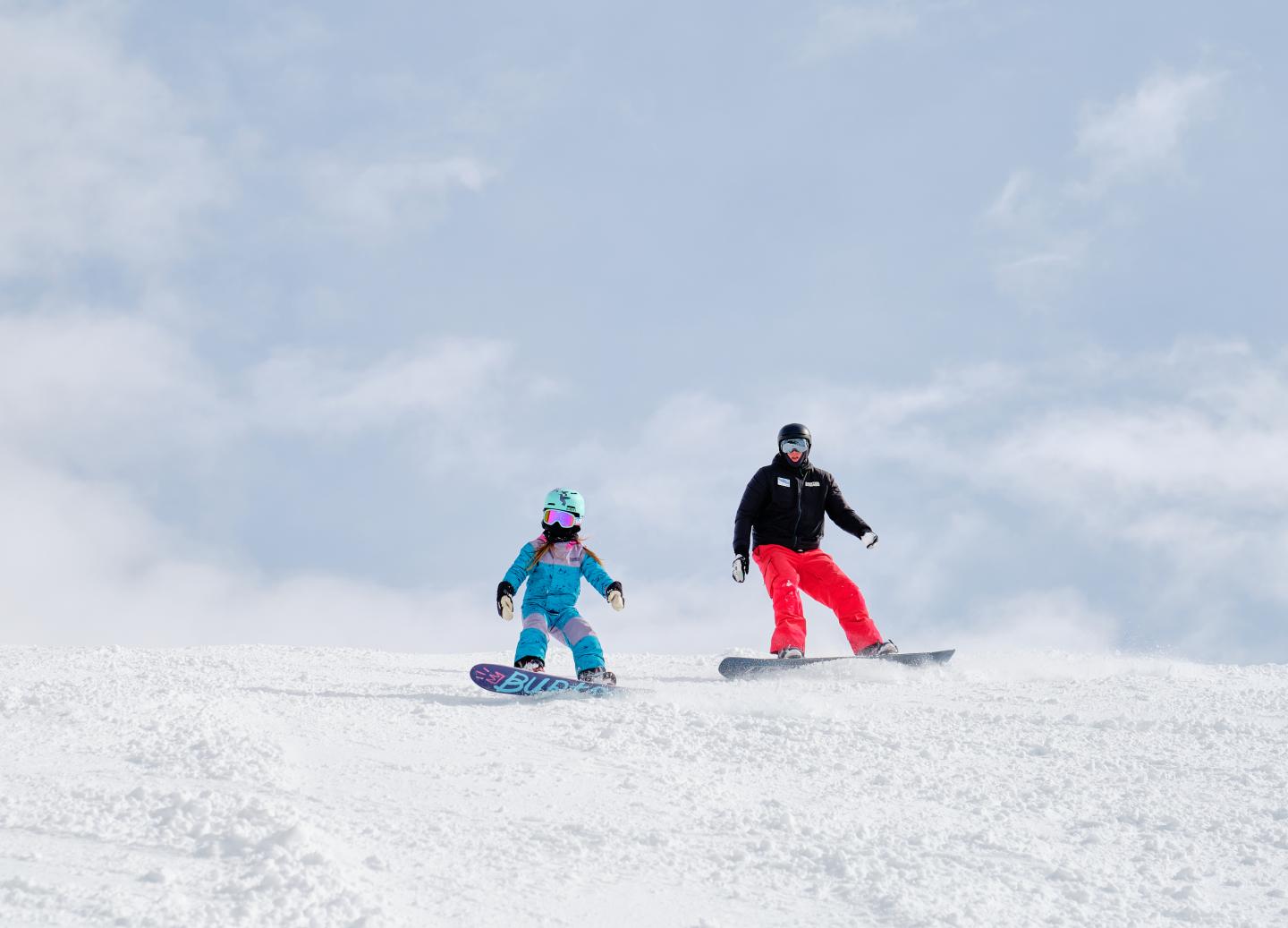 Two snowboarders descending a snowy slope under a cloudy blue sky.