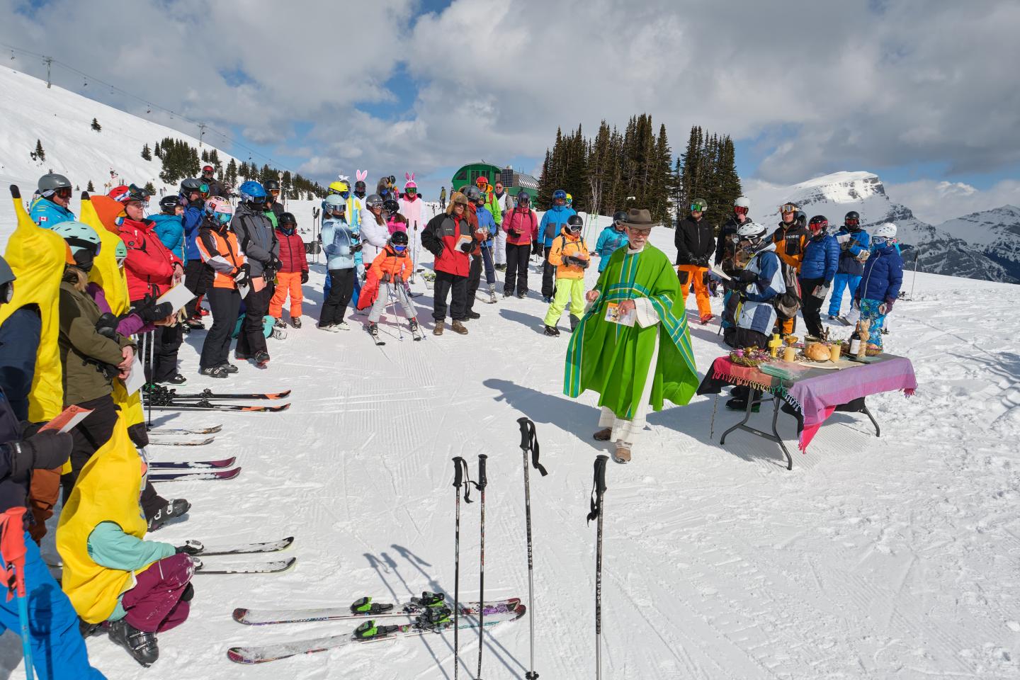 Skiers in colorful gear gather on a snowy mountain under cloudy skies.