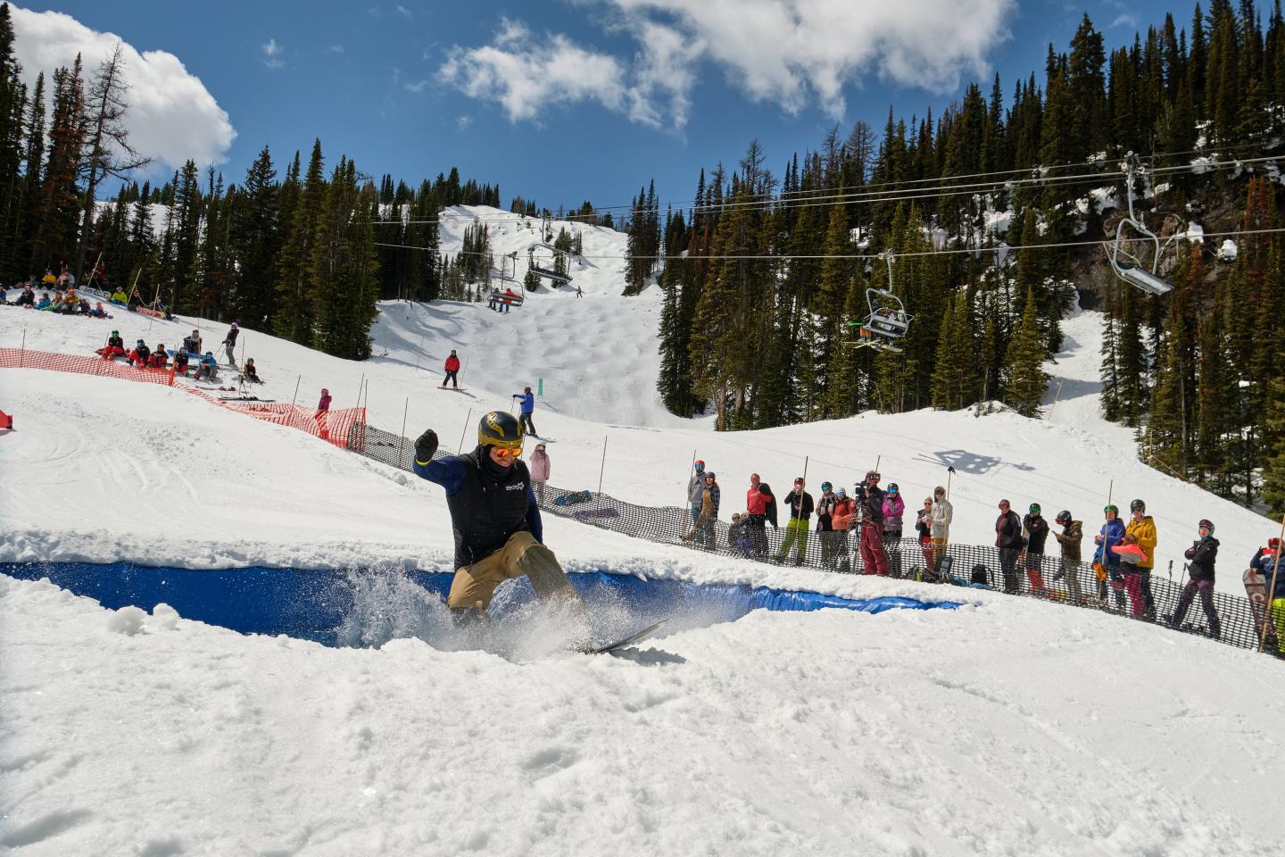 Snowboarder jumping over a snowy ridge, with spectators and pine-covered mountains behind.