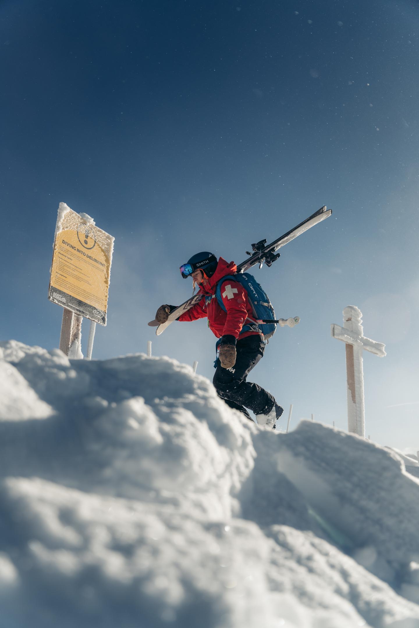 Skier in red jacket climbs a snowy ridge carrying skis under a blue sky.