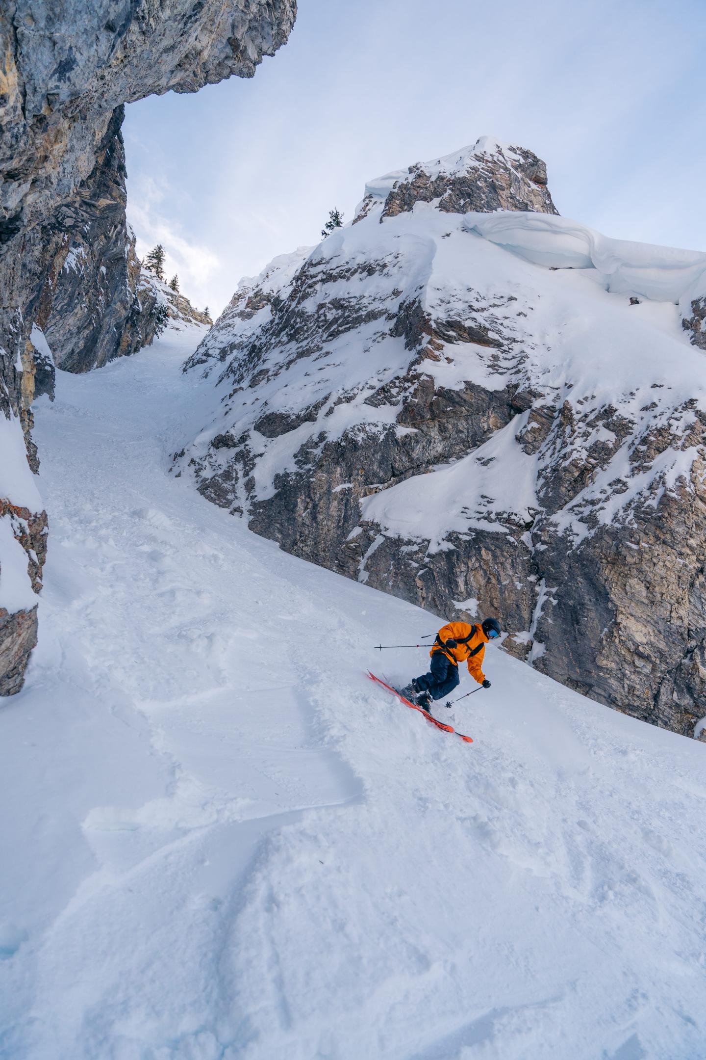 Skier in an orange jacket carving down a steep snowy mountain chute.