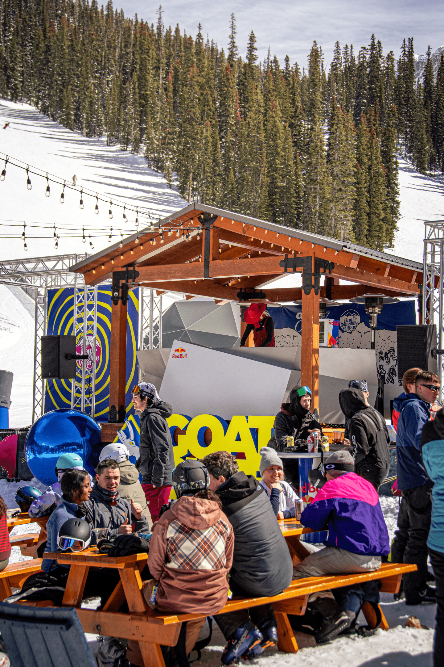 Skiers gather at a colorful outdoor lodge patio beneath snowy pine-covered slopes.