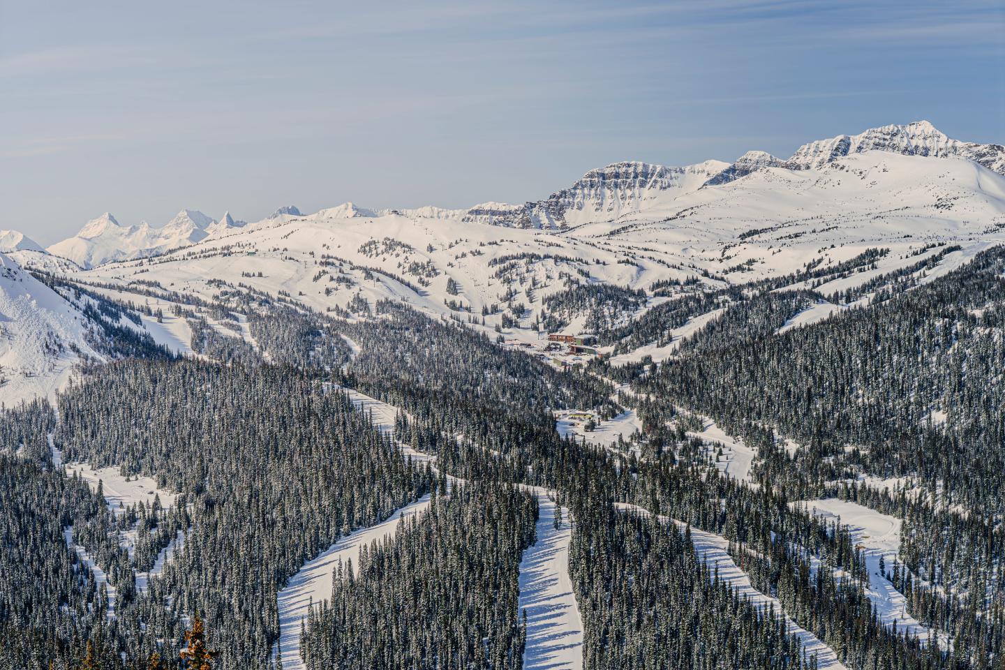 Snow-covered mountain valley with evergreen forests and ski runs under a pale blue sky.