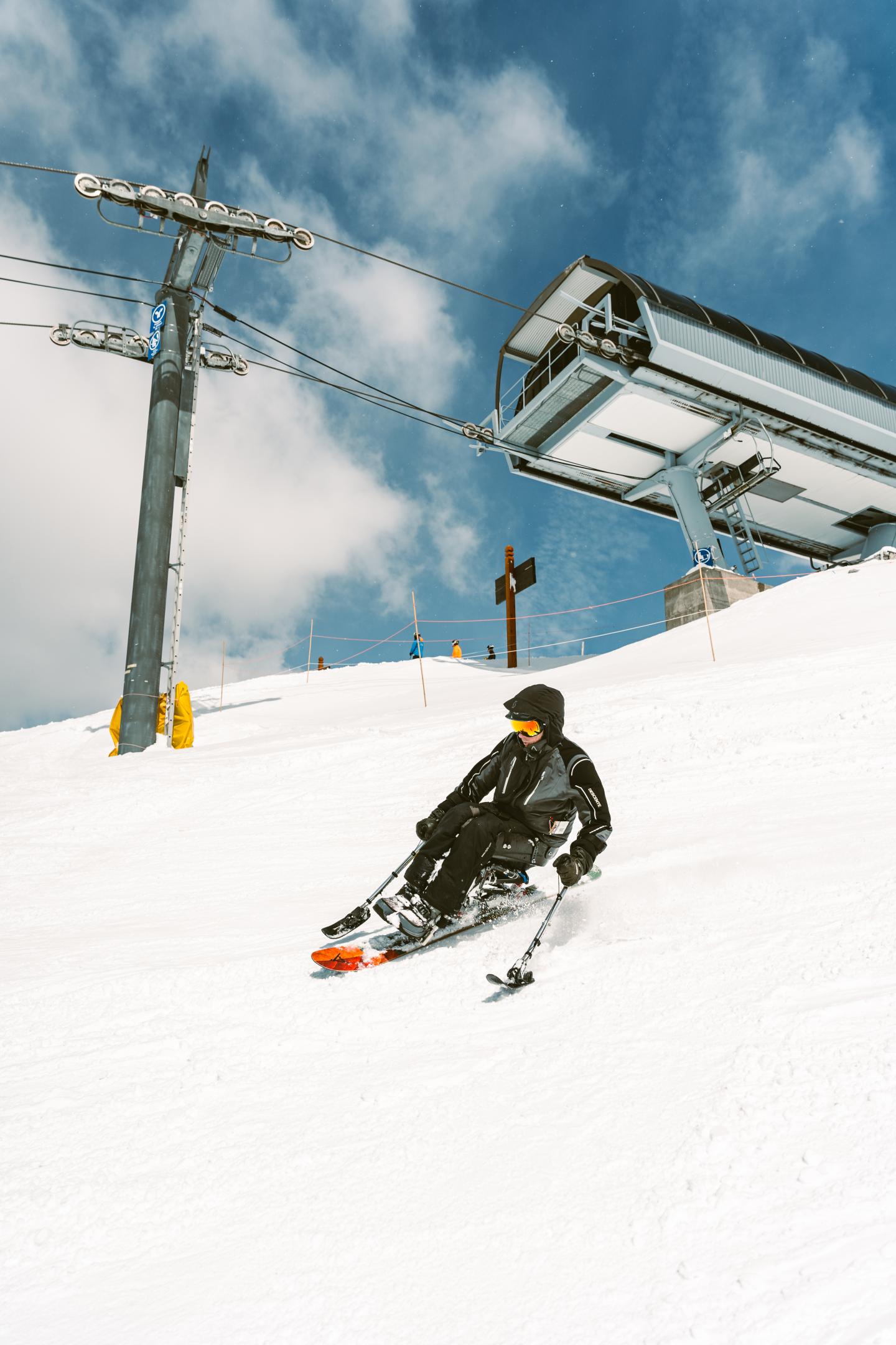 Sit skier carving down a snowy slope beneath a chairlift and blue sky.