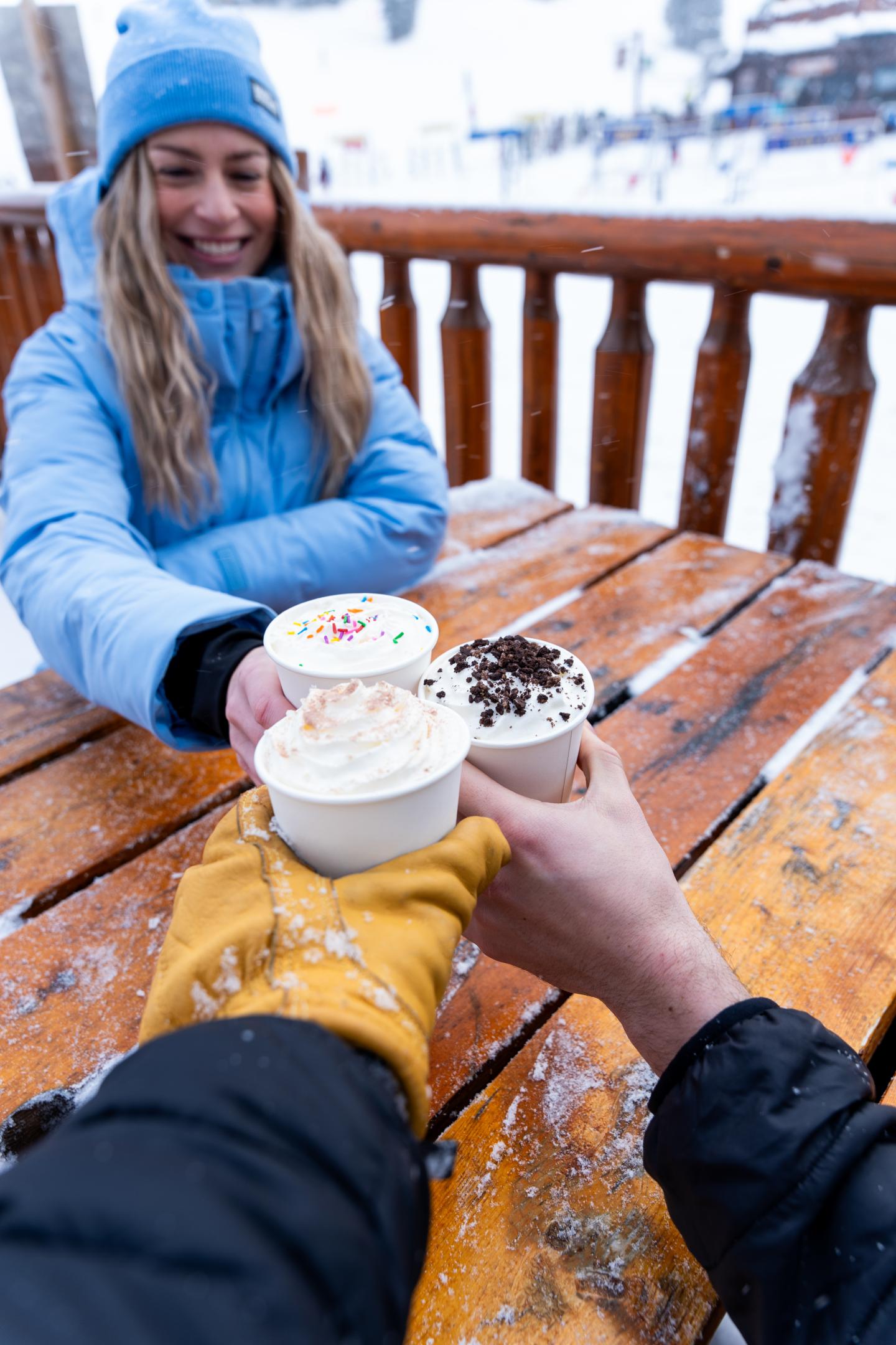 Woman and companion toast hot drinks at a snowy outdoor table.