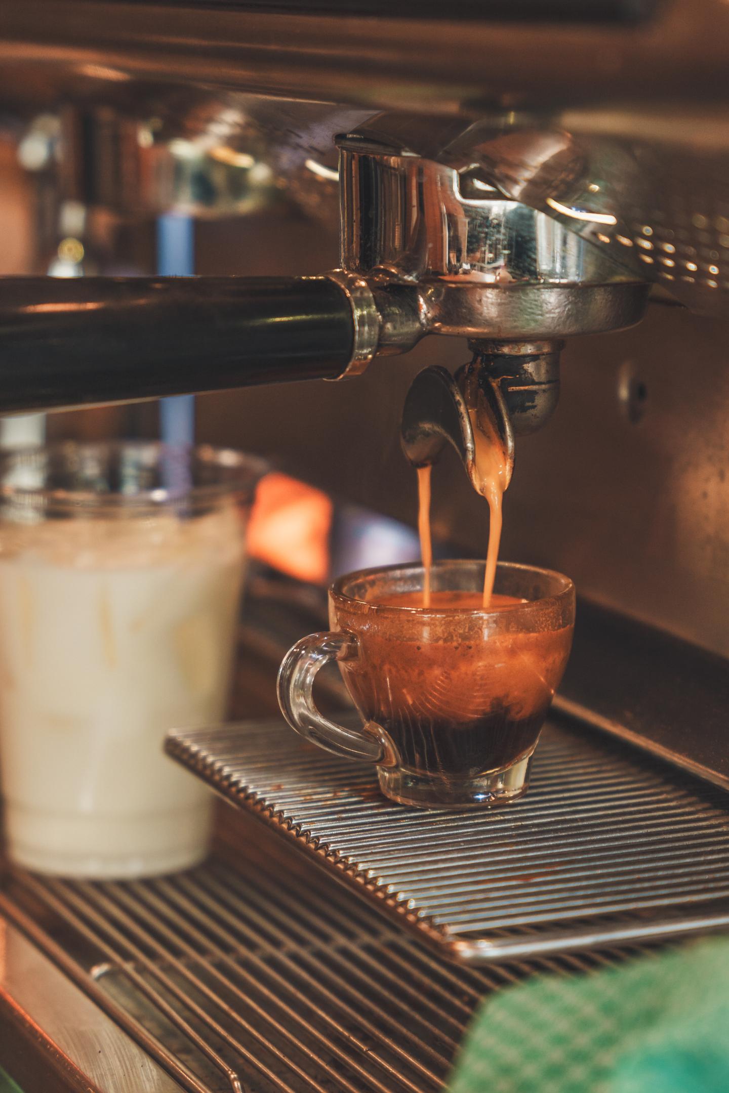 Espresso pours from a machine into a small glass beside a cup of milk.