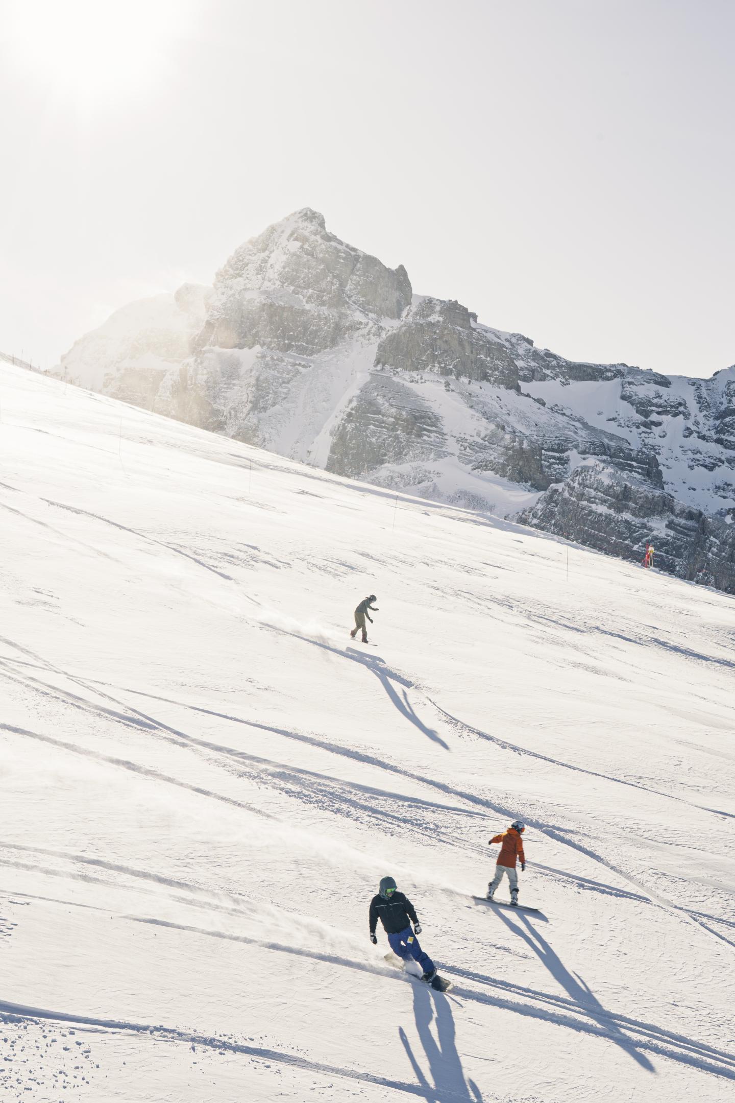 Three skiers descend a snowy slope beneath rugged mountain peaks.