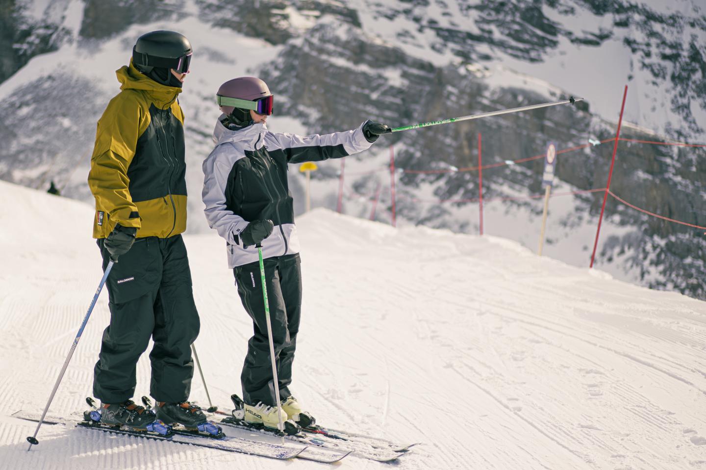 Two skiers on a snowy mountain slope, one pointing downhill.
