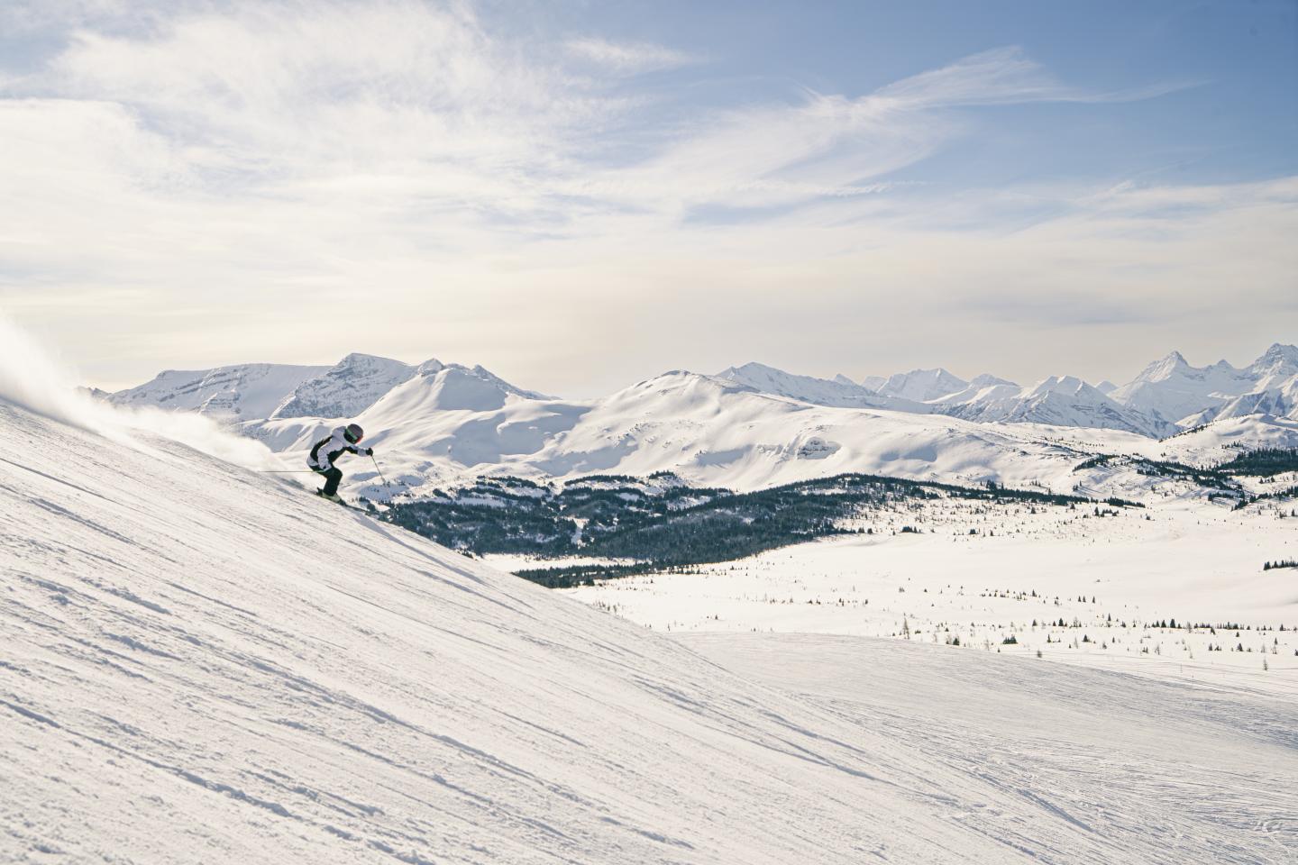 Skier descending a snowy mountain slope with rugged peaks in the distance.