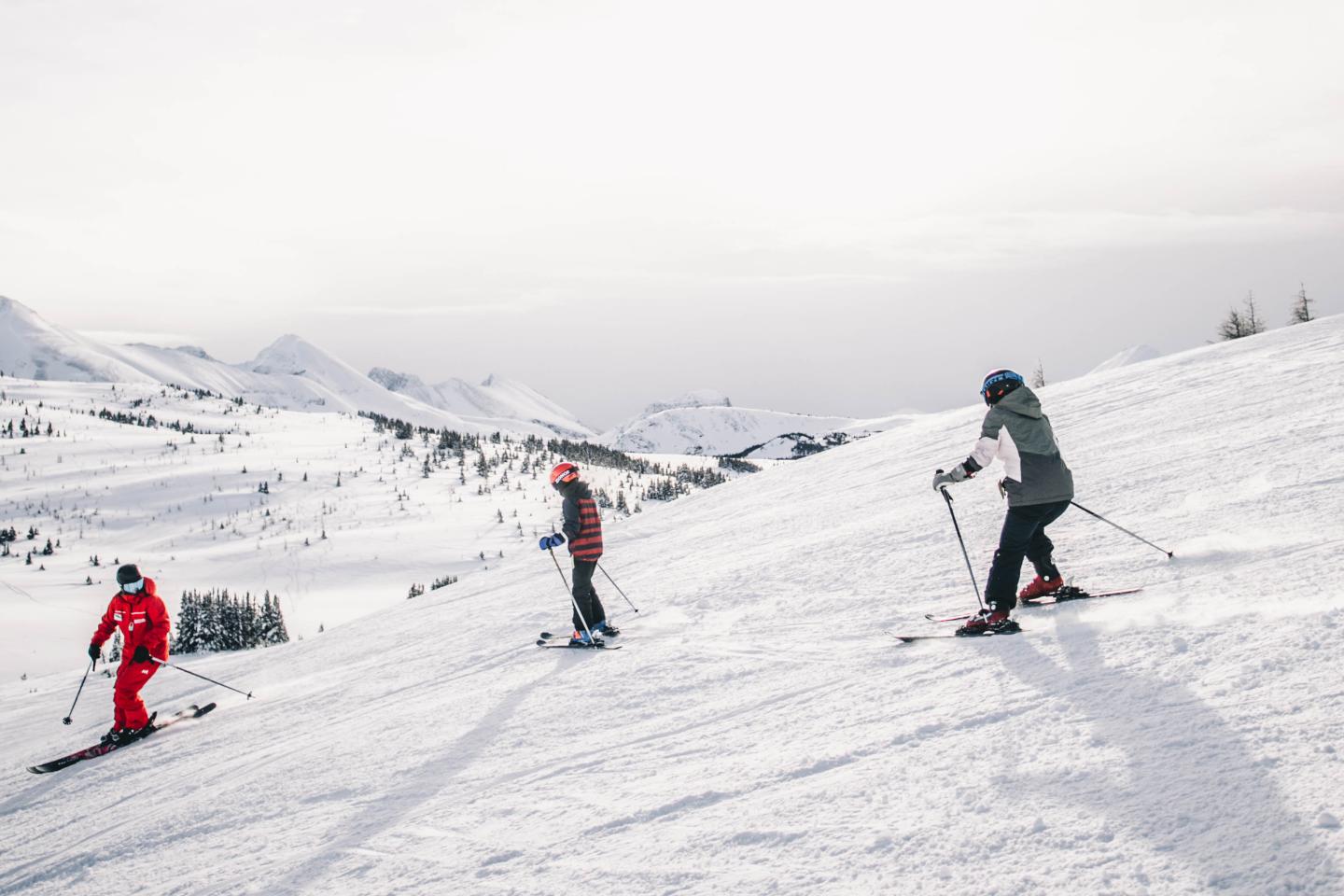 Three skiers descend a snowy mountain slope under a cloudy sky.