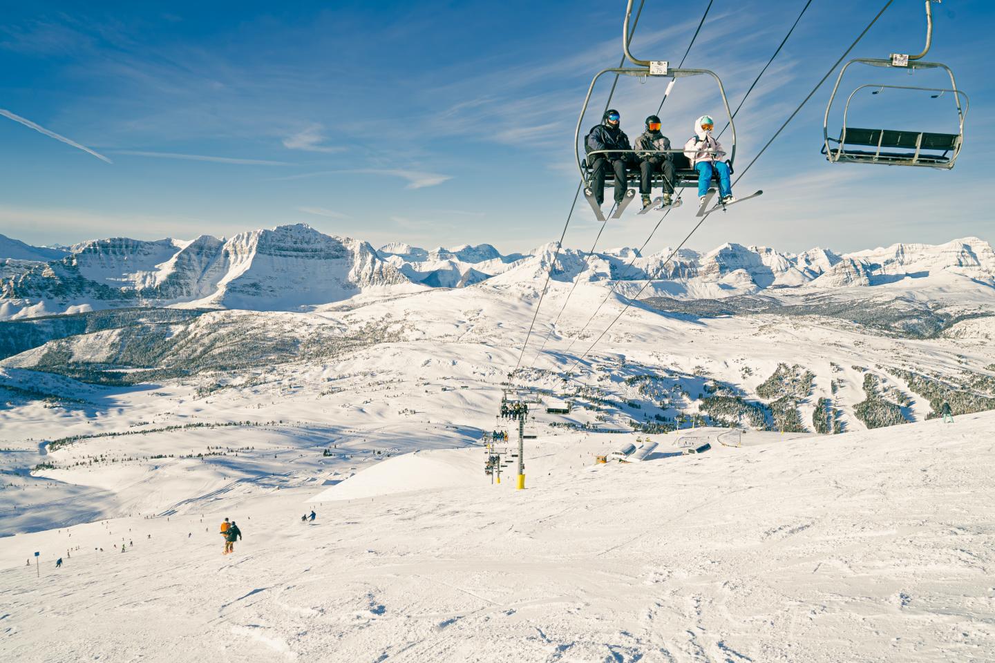 Skiers ride chairlifts above a snowy mountain slope under a blue sky.