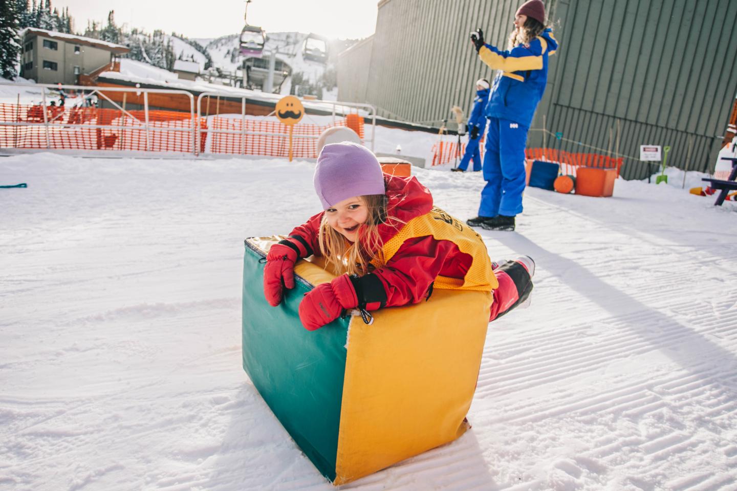 Child in a colorful toy car on snow while an adult stands nearby.