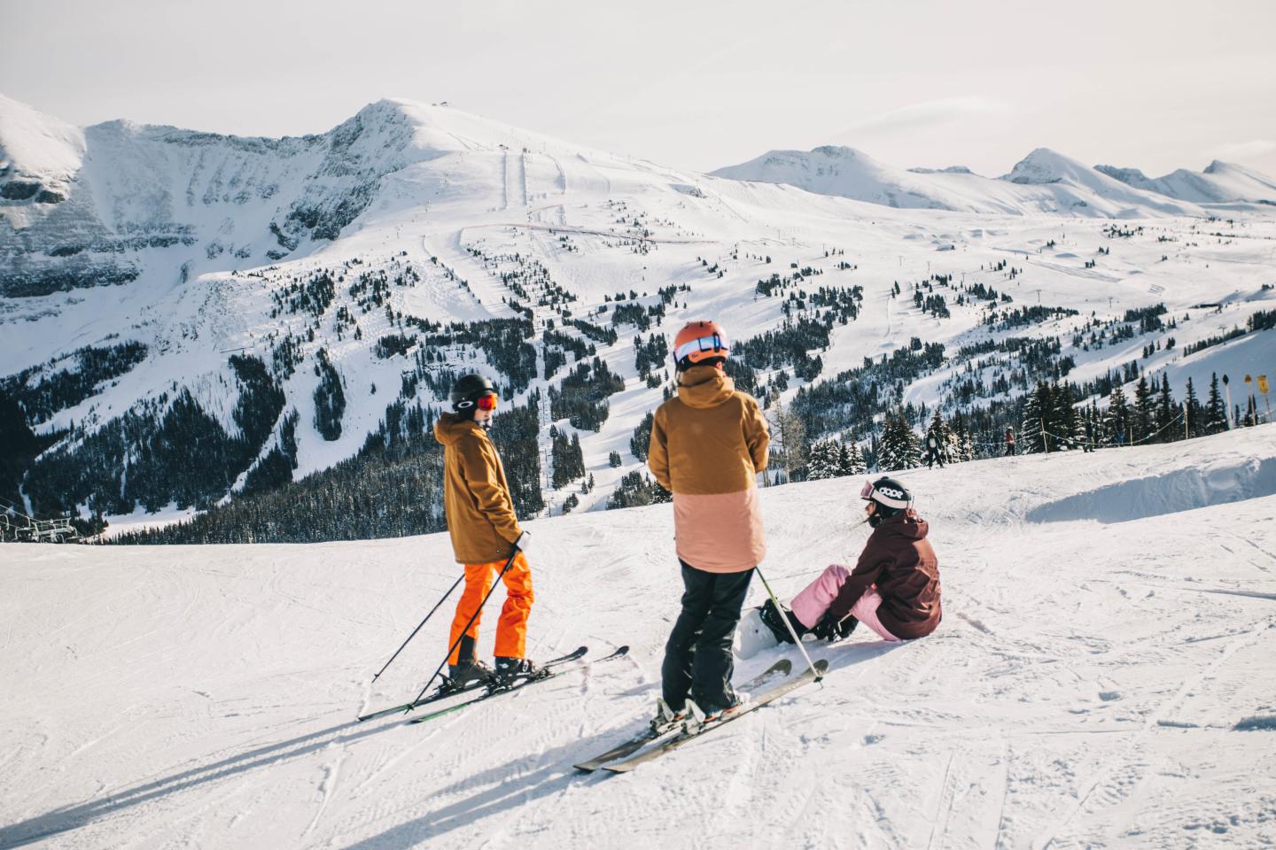 Three skiers pause on a snowy mountain slope beneath rugged alpine peaks.
