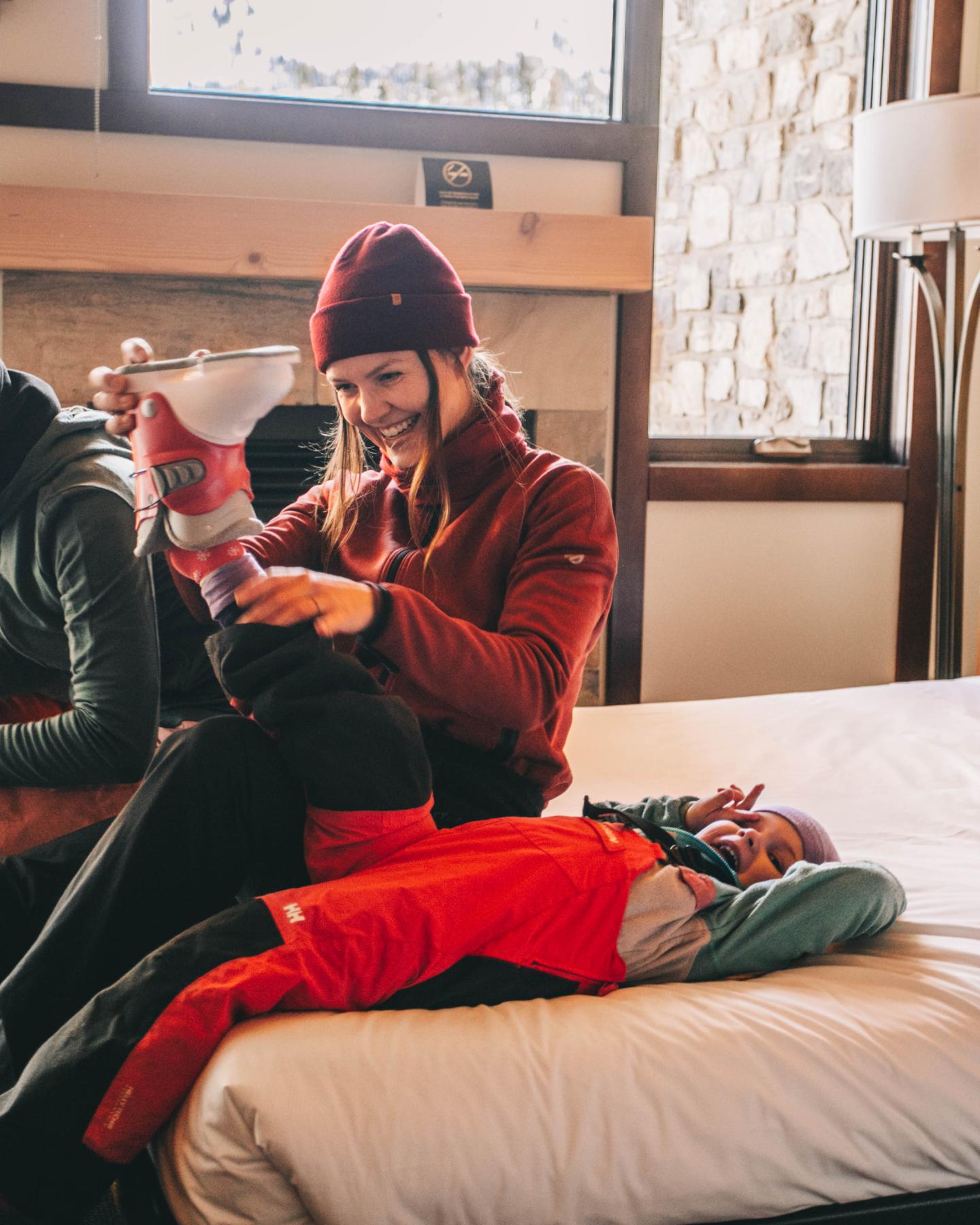 Man in a beanie checks his phone beside a bundled child on a bed.