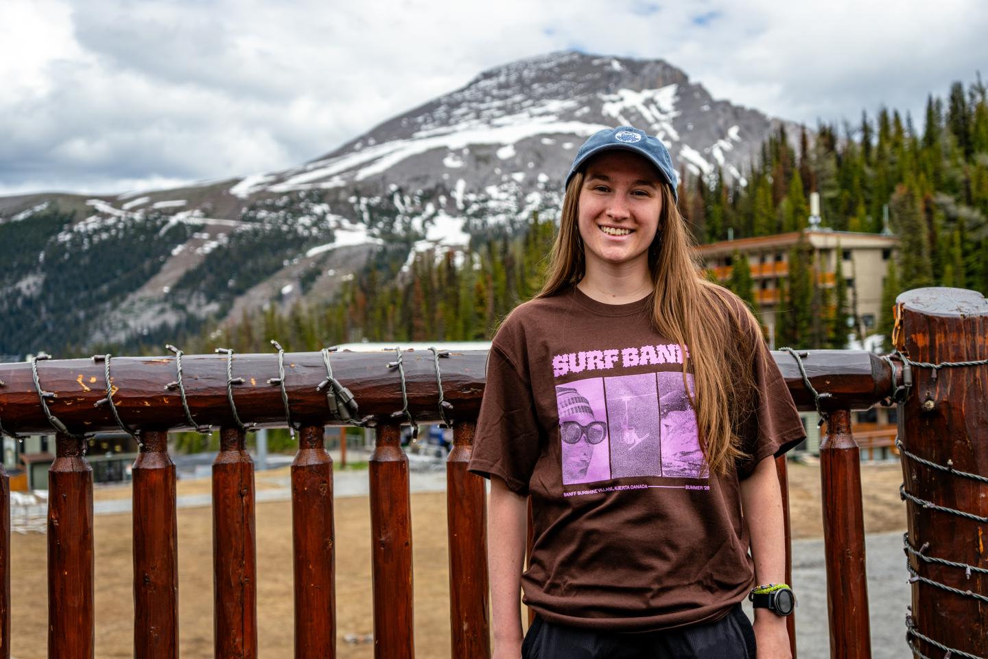Smiling woman in a cap at a wooden fence with snowy mountains behind.