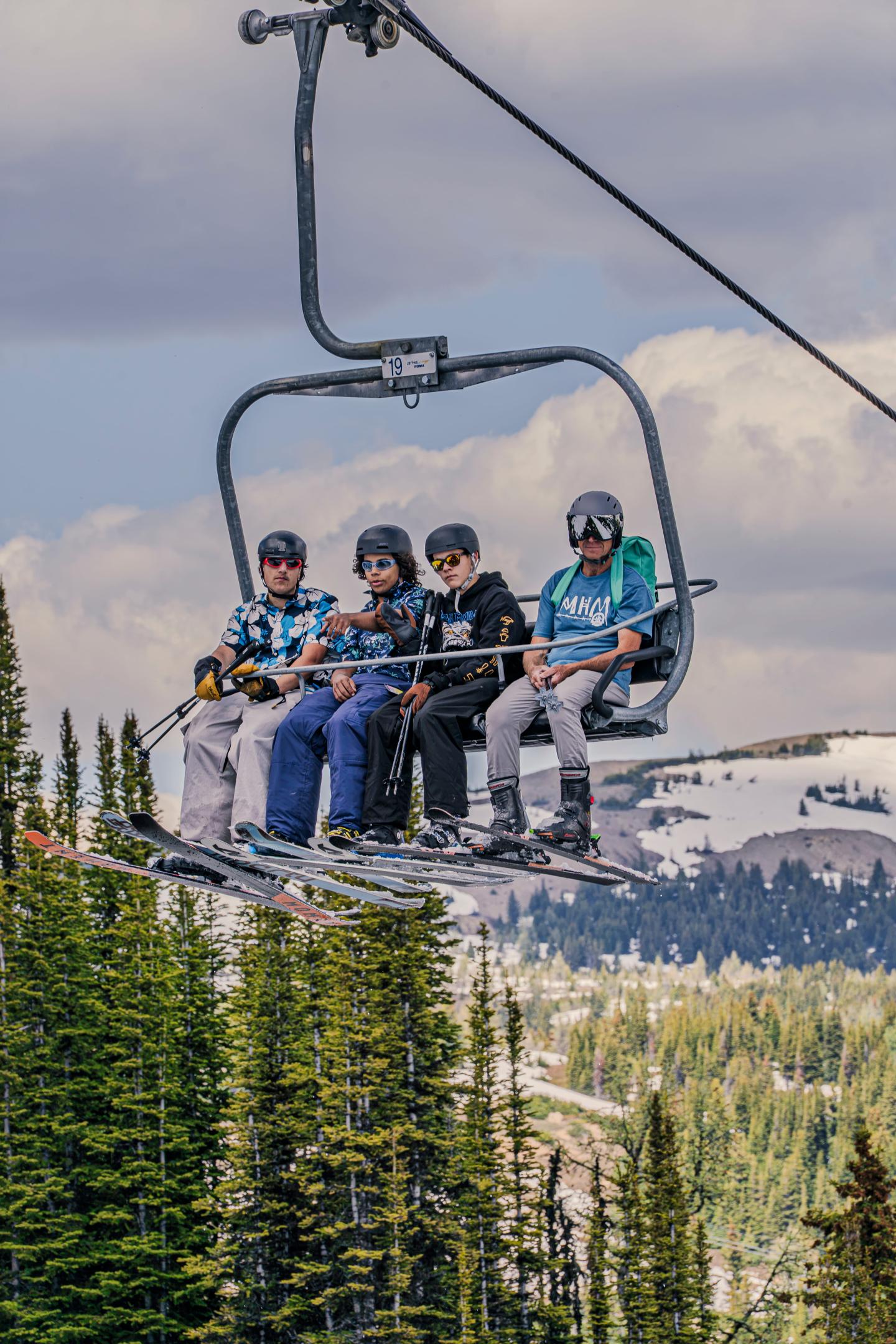 Four skiers riding a chairlift above snow-covered mountains and evergreen trees.