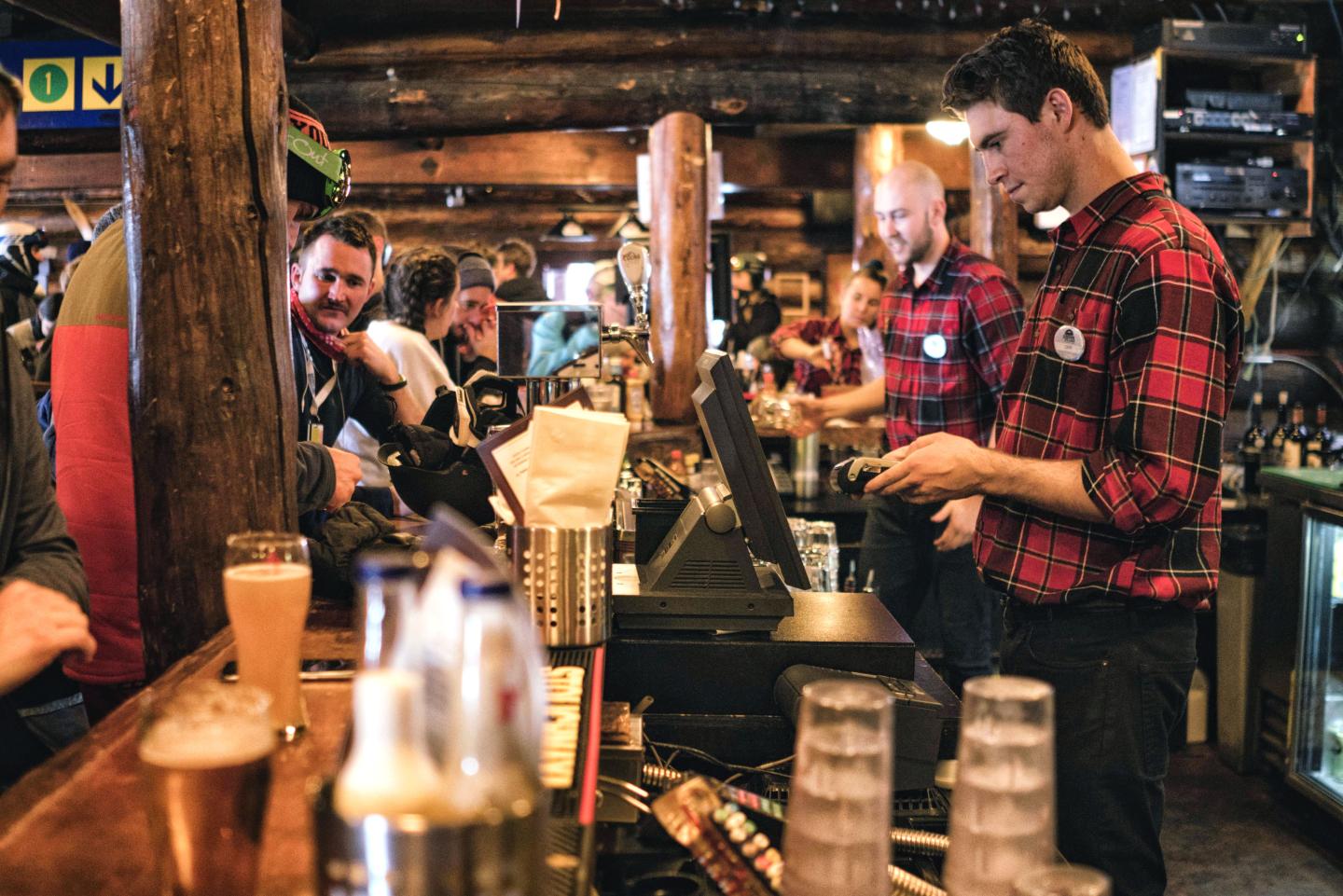 Barista in red plaid shirt preparing coffee behind a busy café counter.