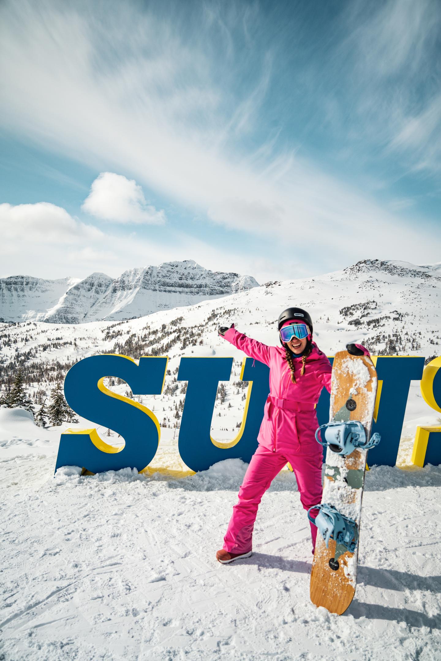 Snowboarder in pink gear posing with board before snowy mountains
