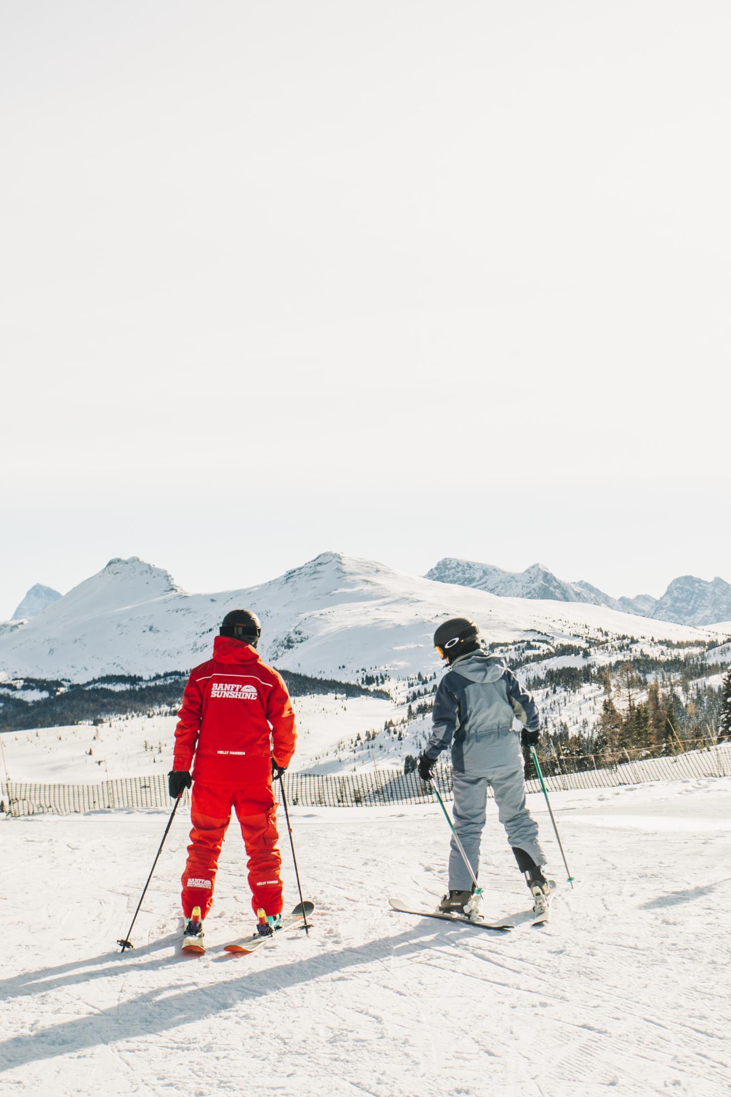 Two skiers in red and gray outfits pause on a snowy mountain slope.