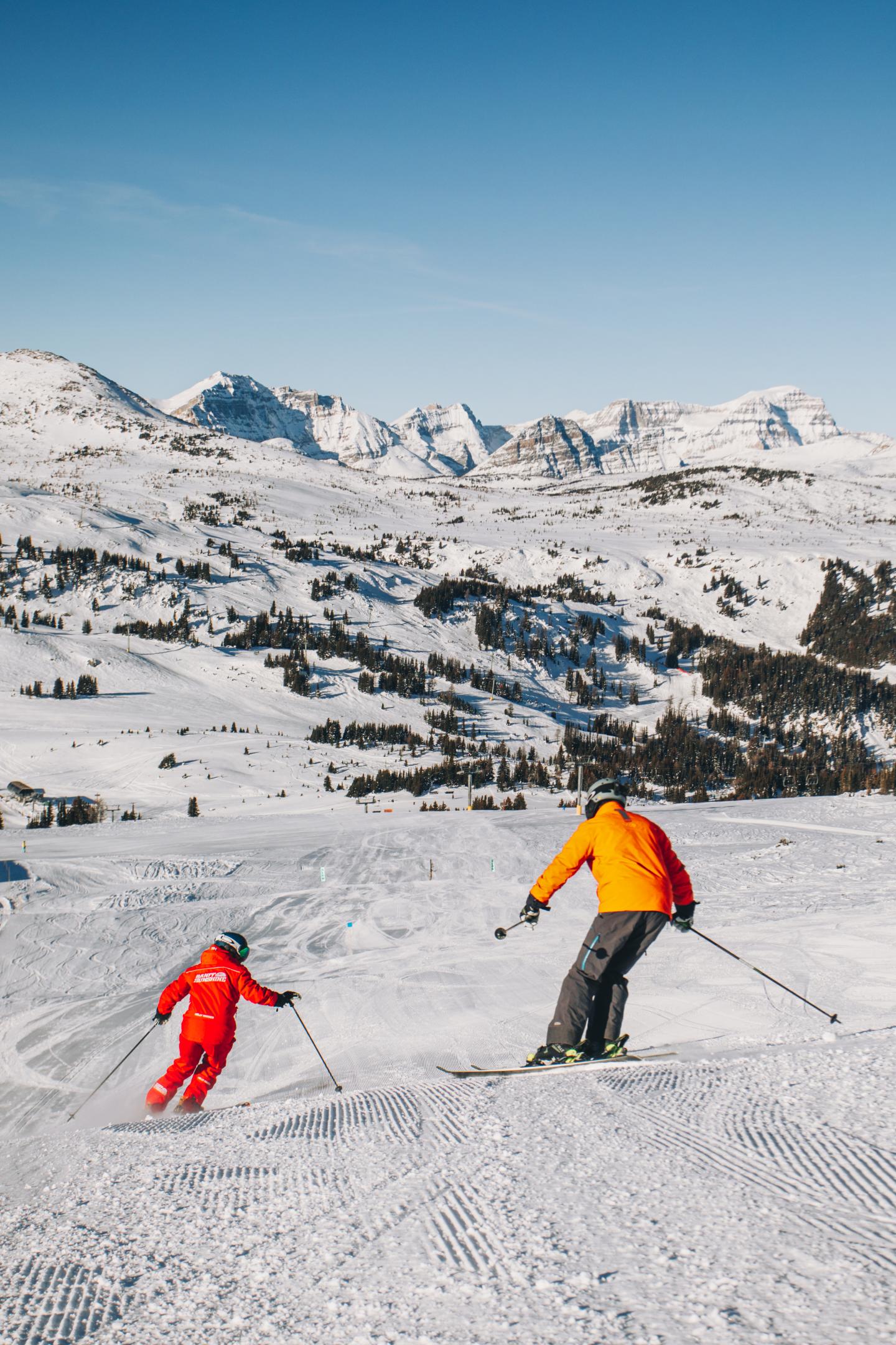 Two skiers descend a snowy slope beneath rugged, sunlit mountains.