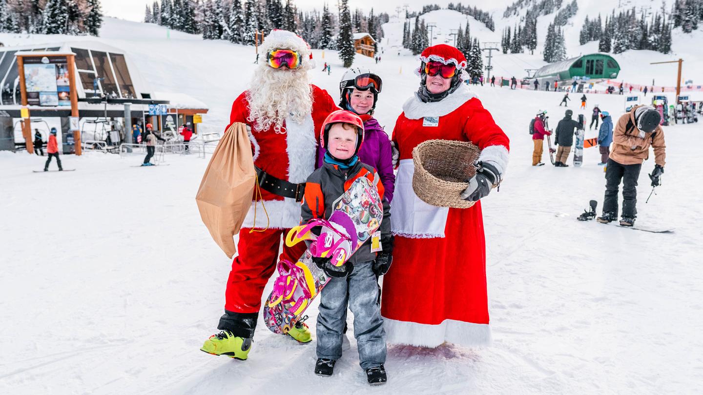 Three people in Santa costumes pose with a child on a snowy ski slope.