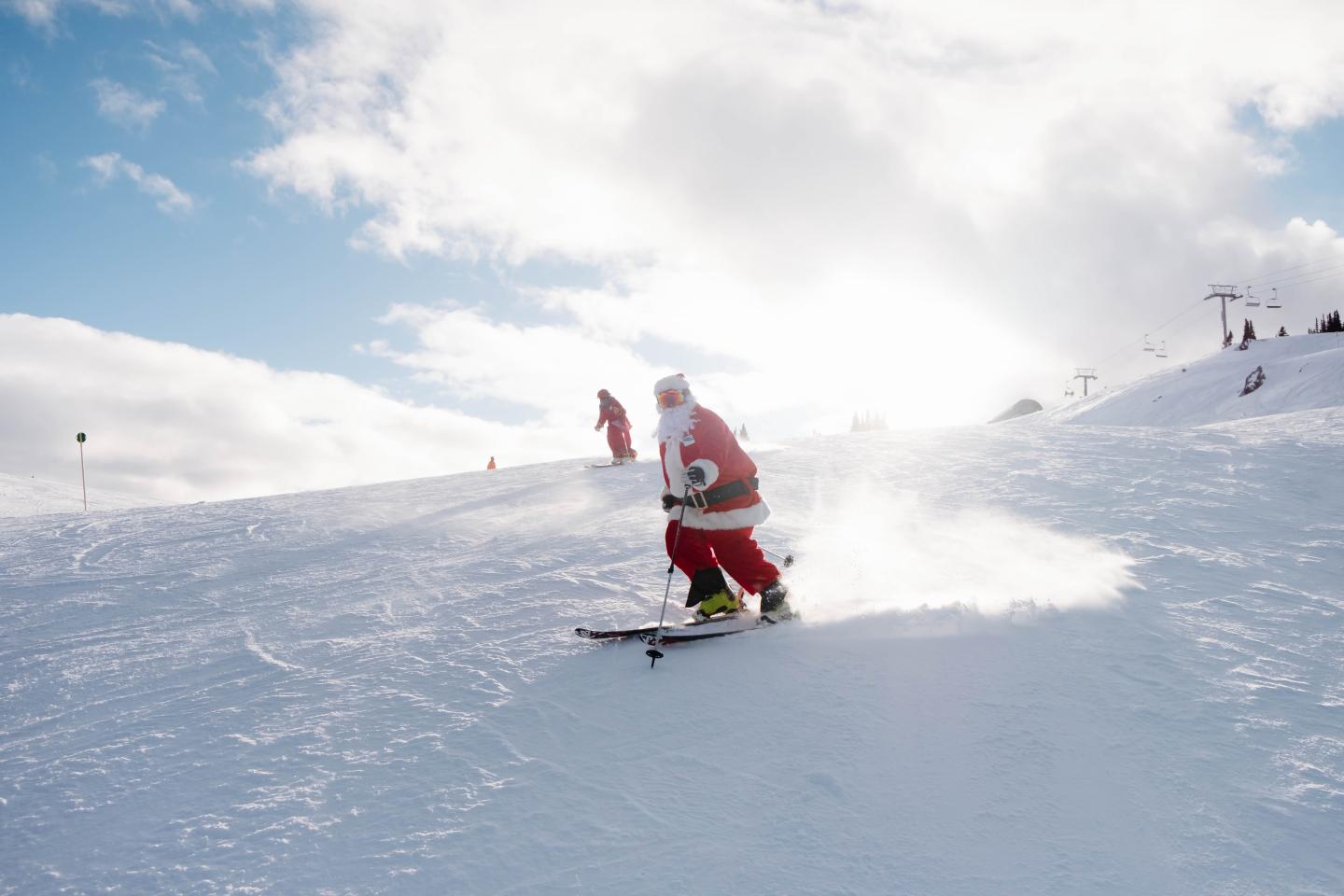 Santa Claus skiing downhill on a snowy mountain under a bright sky.