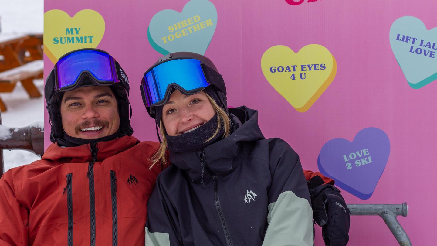 Two bundled-up skiers smile before a colorful heart-shaped backdrop.