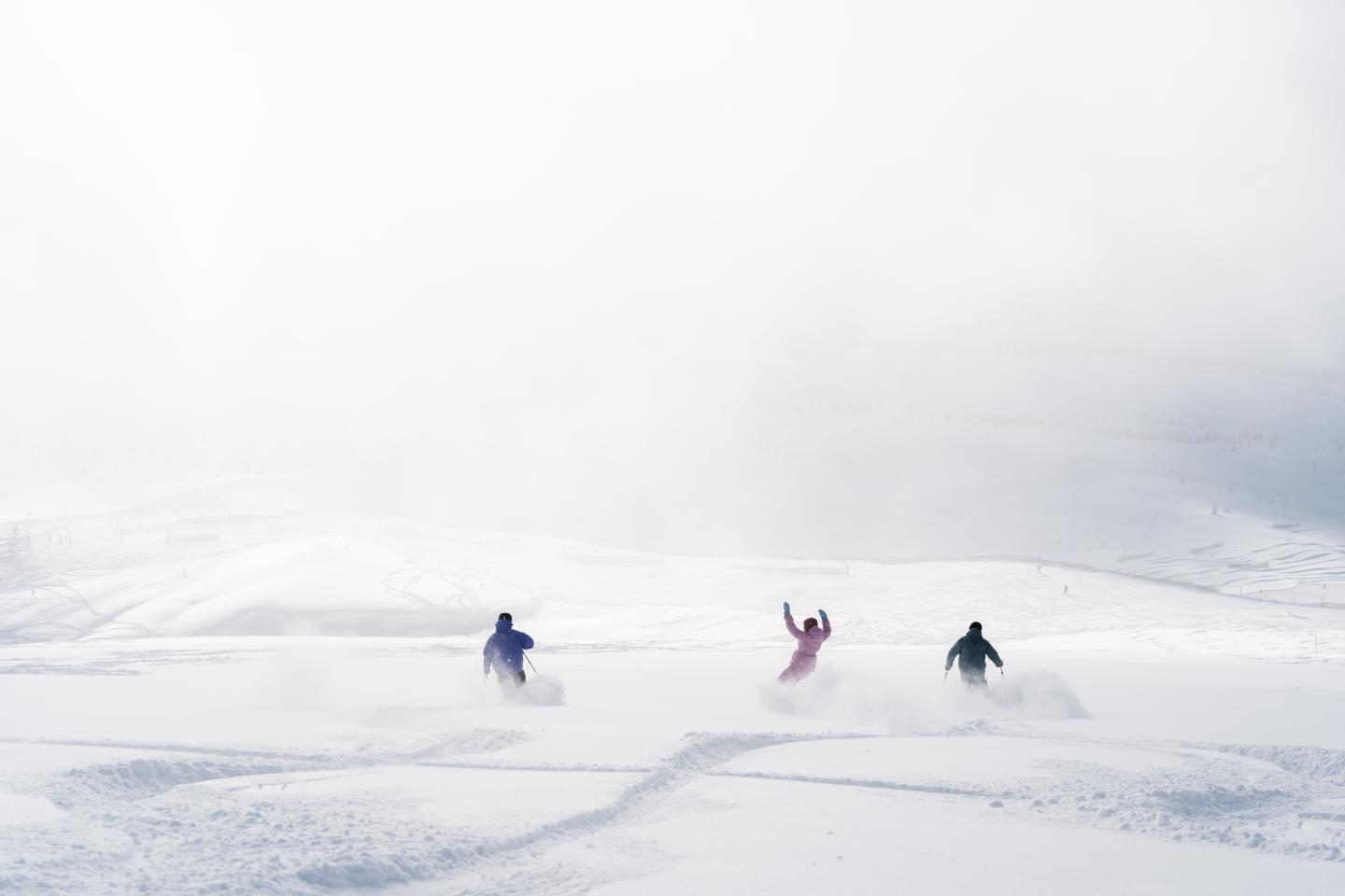 Three skiers traverse a snowy mountain slope beneath a cloudy sky.