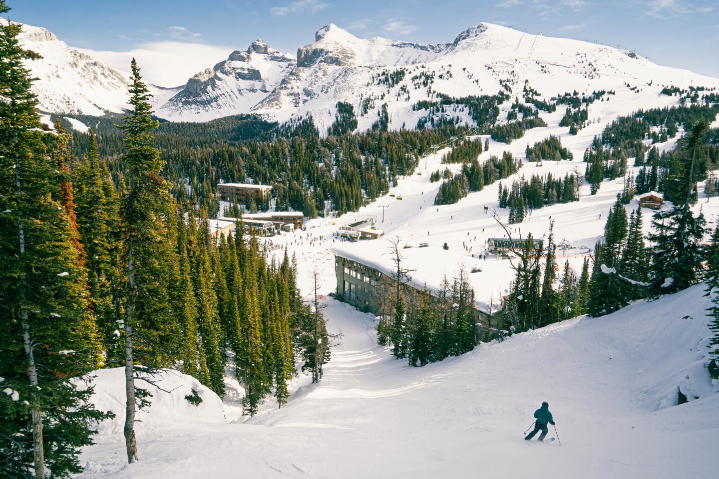 Skier descending a snowy slope beneath rugged mountain peaks and pine trees.