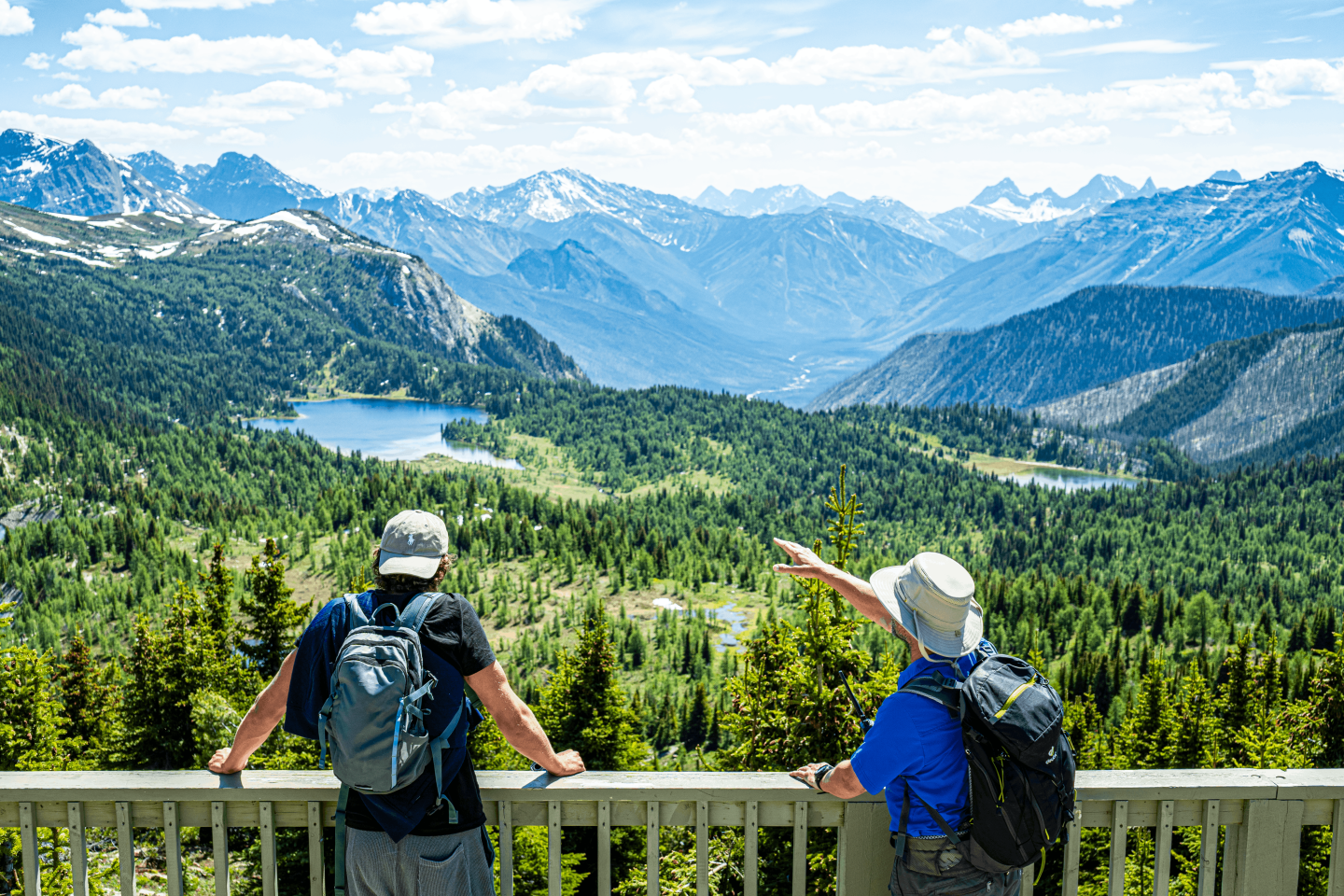 Two hikers overlook a forested mountain valley beneath snow-capped peaks.