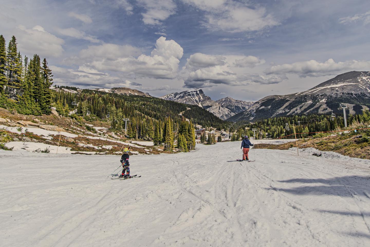 Two hikers cross a snowy alpine valley beneath rugged mountains and cloudy skies.
