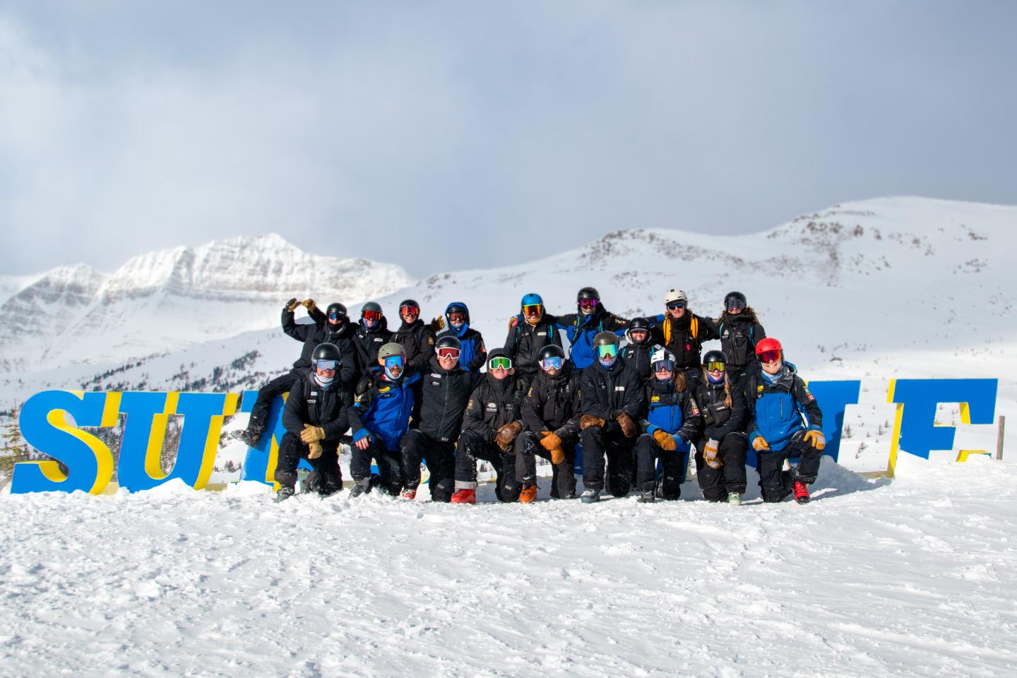Group of skiers in dark gear posing on snowy mountains beside colorful letters.