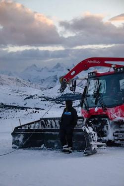 Red snow groomer with driver parked on a snowy mountain slope at dusk.