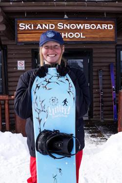 Smiling skier in blue bib overalls holding a helmet outside ski school.