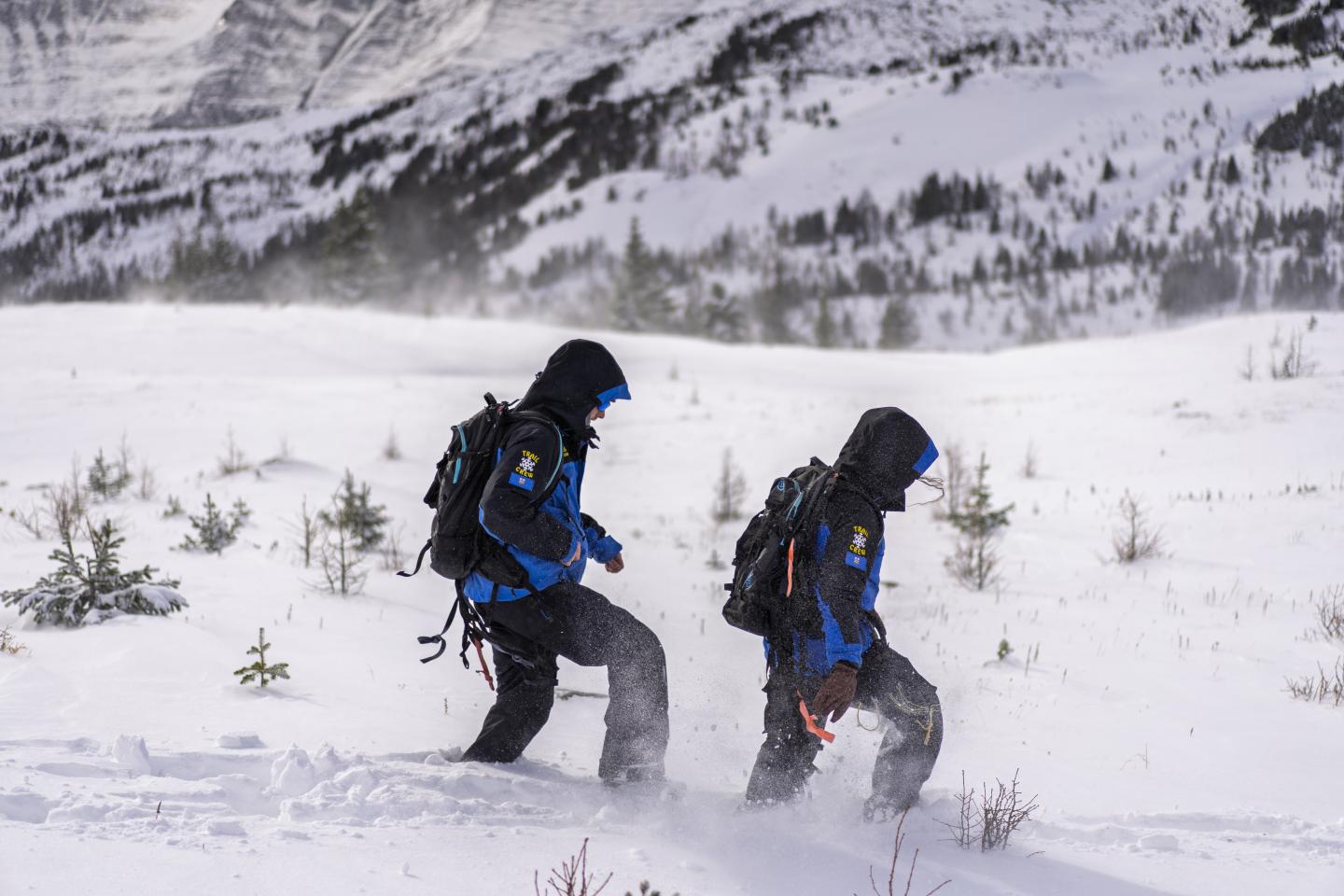 Two hikers in dark winter gear walk through deep snow beneath snowy mountains.