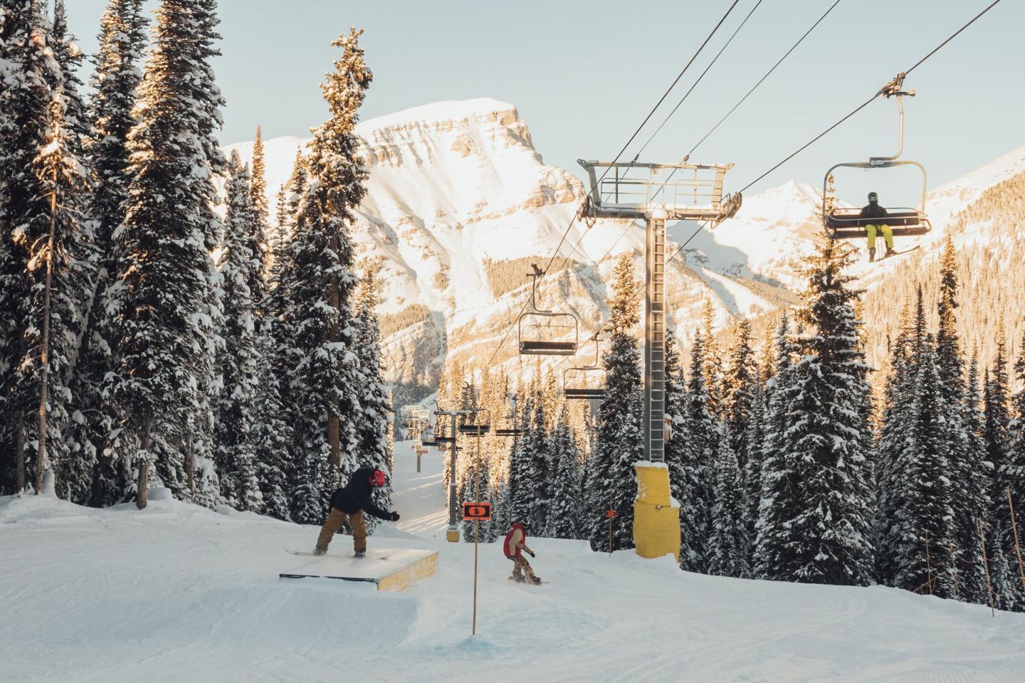 Skier on a snowy chairlift trail framed by pine trees and sunlit mountains